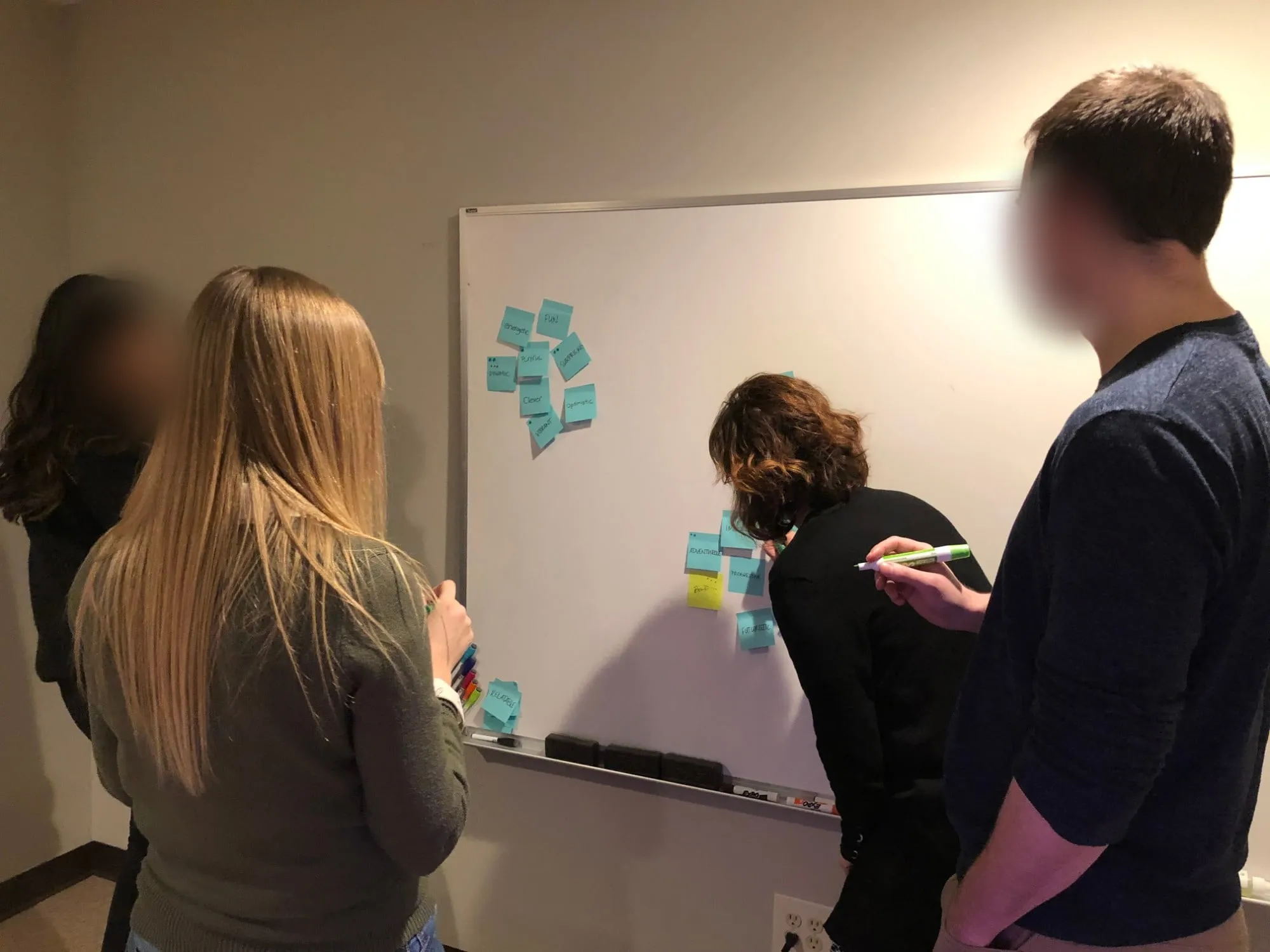 Four people at a whiteboard sorting light blue sticky notes of candidate brand words, among them energetic, fun, playful, surprising, clever, optimistic, adventurous, imaginative, progressive, futuristic and bold, with dot votes on some. Faces are blurred.
