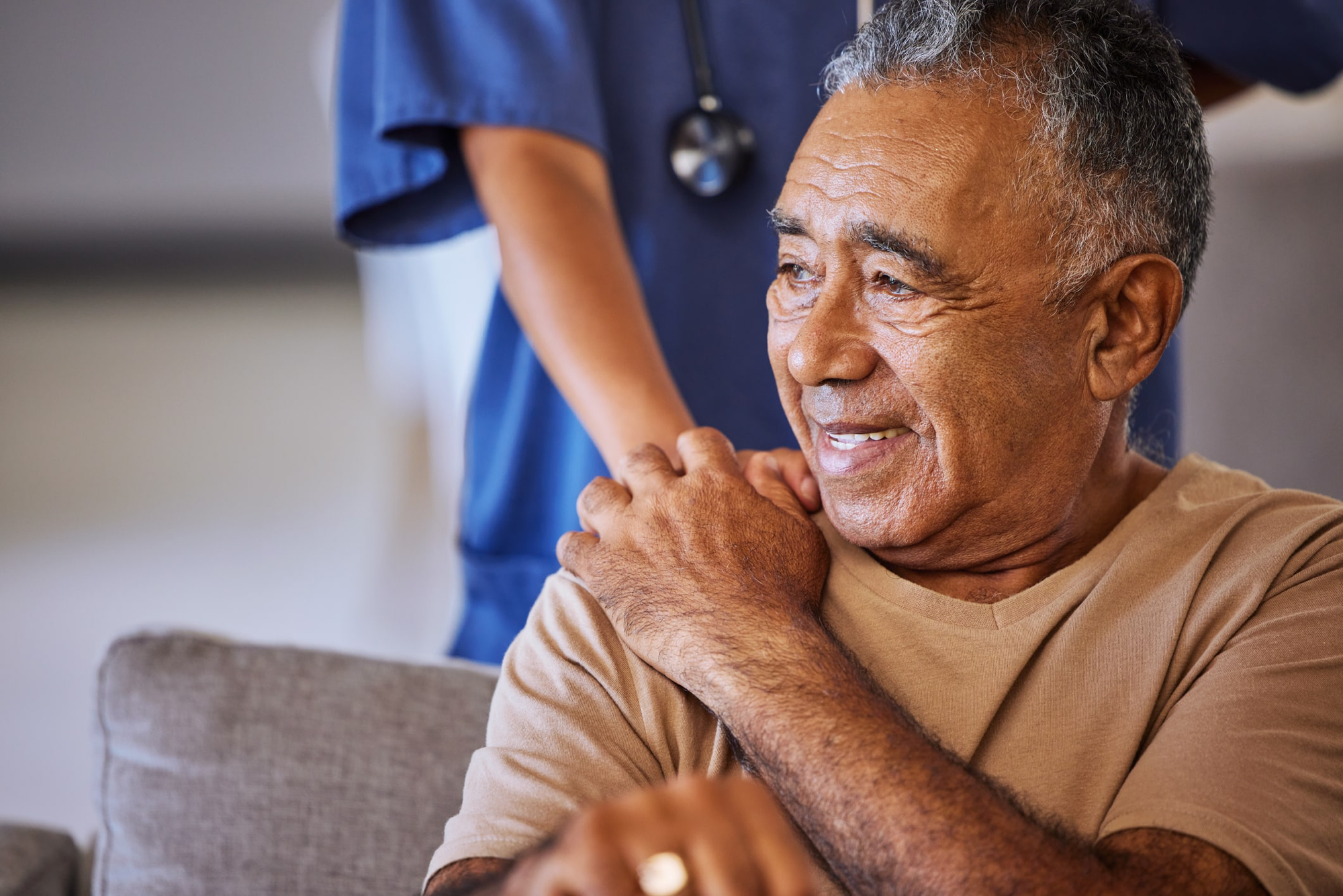 An older man seated on a sofa, smiling, resting his hand over the hand a clinician in blue scrubs has placed on his shoulder. The clinician is behind him and out of focus, a stethoscope round their neck.