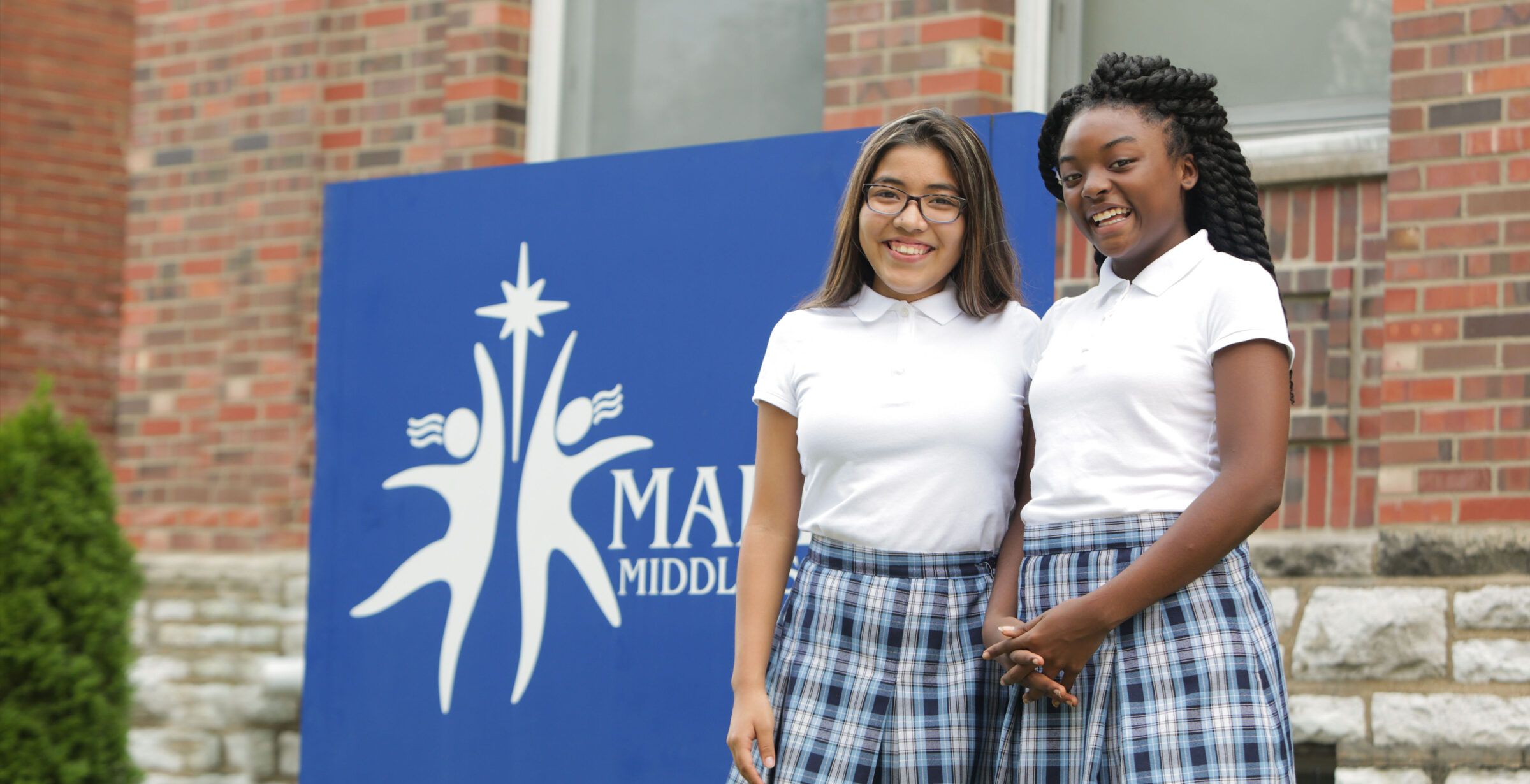 Two Marian students in white uniform polos and plaid skirts standing and smiling in front of the blue Marian Middle School sign, with the school's brick facade behind them