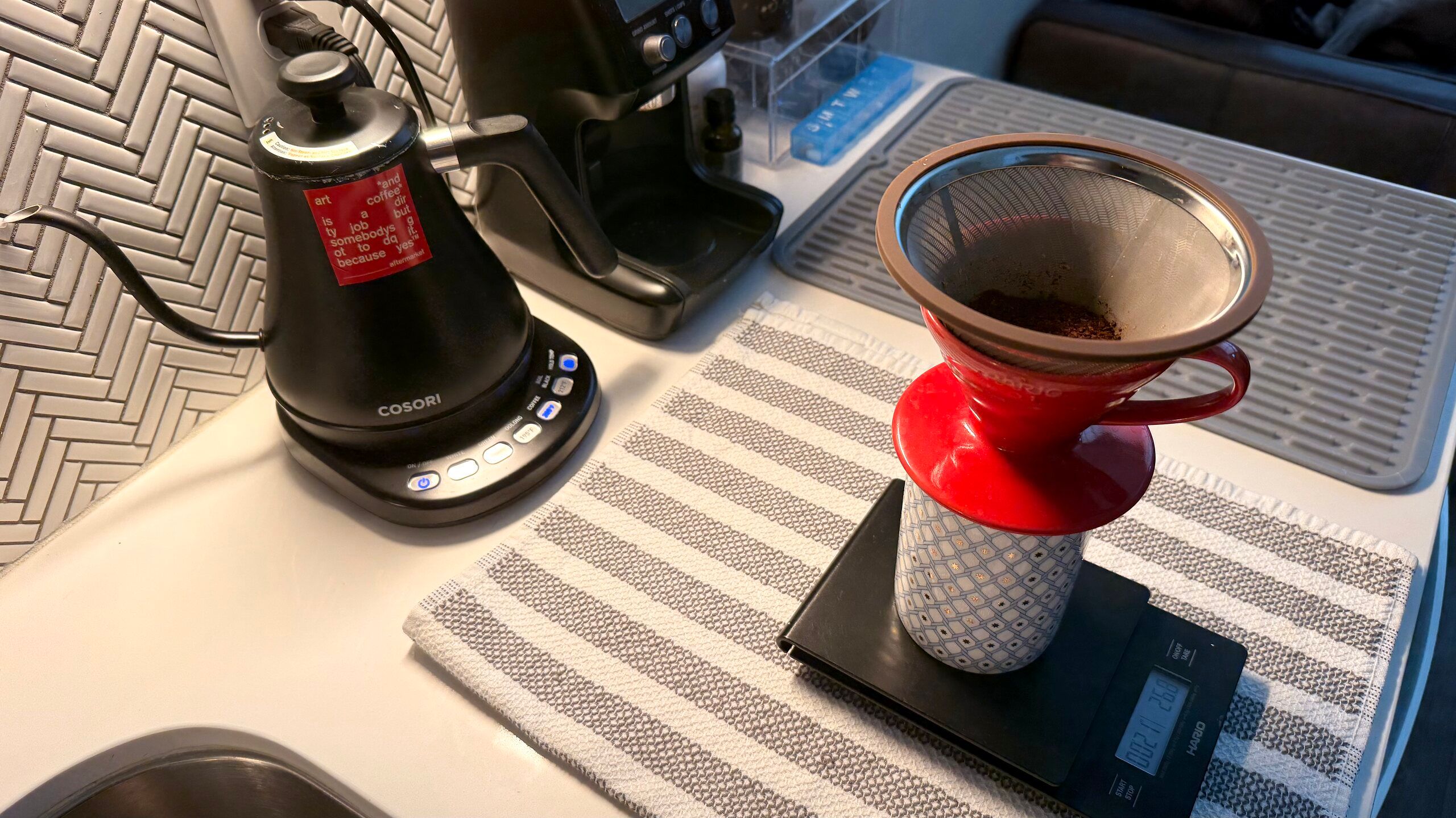 Mid-brew at the pour-over station: a stainless mesh filter loaded with grounds sits in the red ceramic dripper on a patterned cup, the Hario scale reading live beneath it, gooseneck kettle heated at the left