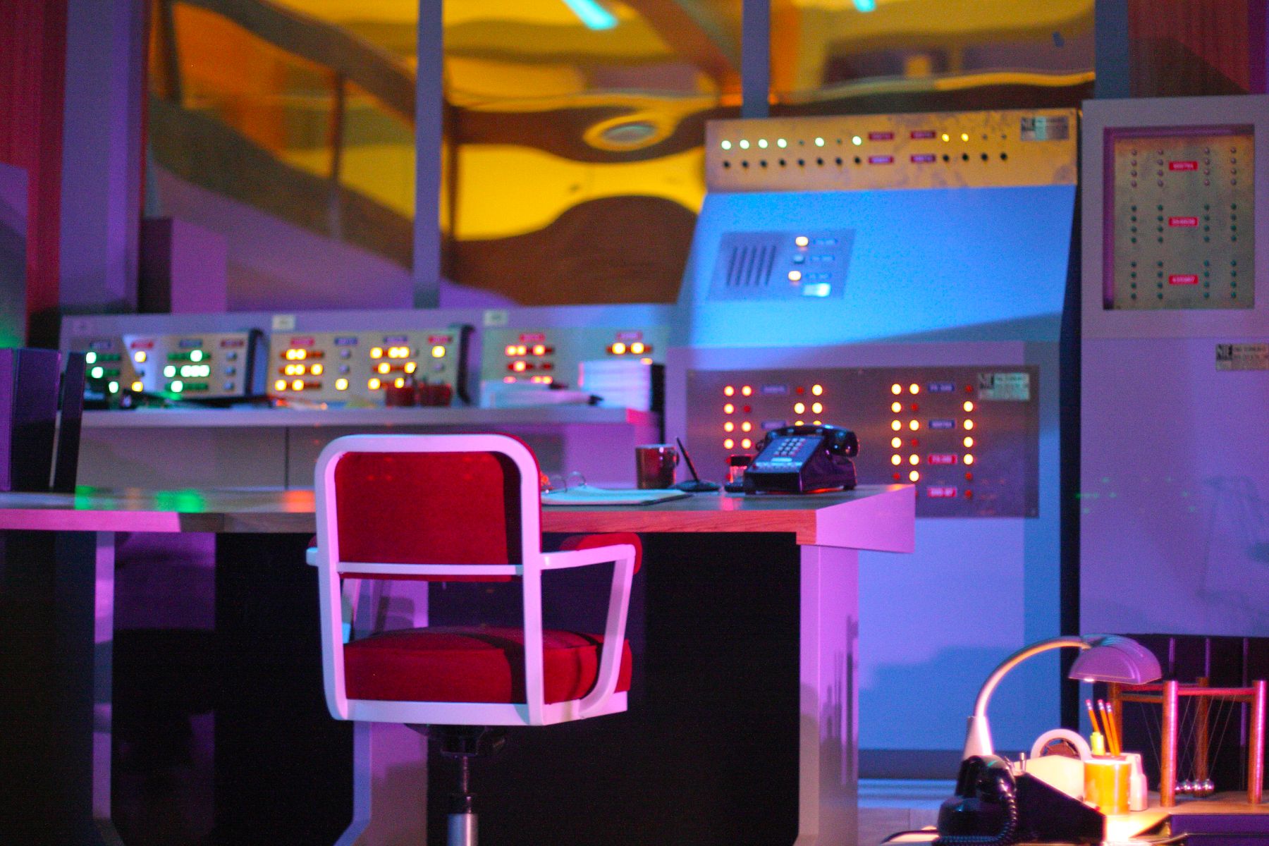 Network operations center bathed in violet and amber light: a workstation of stacked monitors and control consoles with an empty red chair facing them, status equipment and panels lining the wall behind.