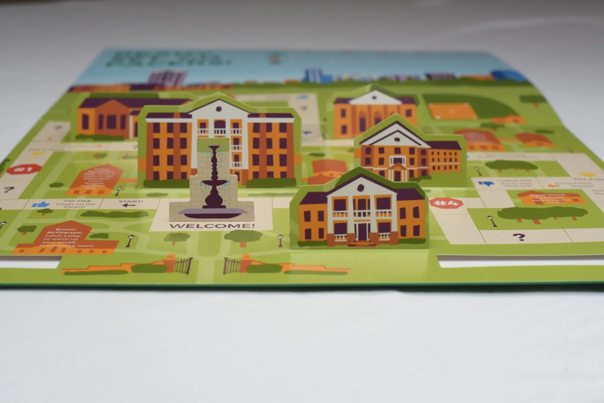 A printed pop-up campus tour map for William Peace University, photographed low across a table: illustrated orange and cream campus buildings and a fountain stand up from a green map, with a WELCOME! square at the start of a game-board path, numbered stops and short captions beside each building.